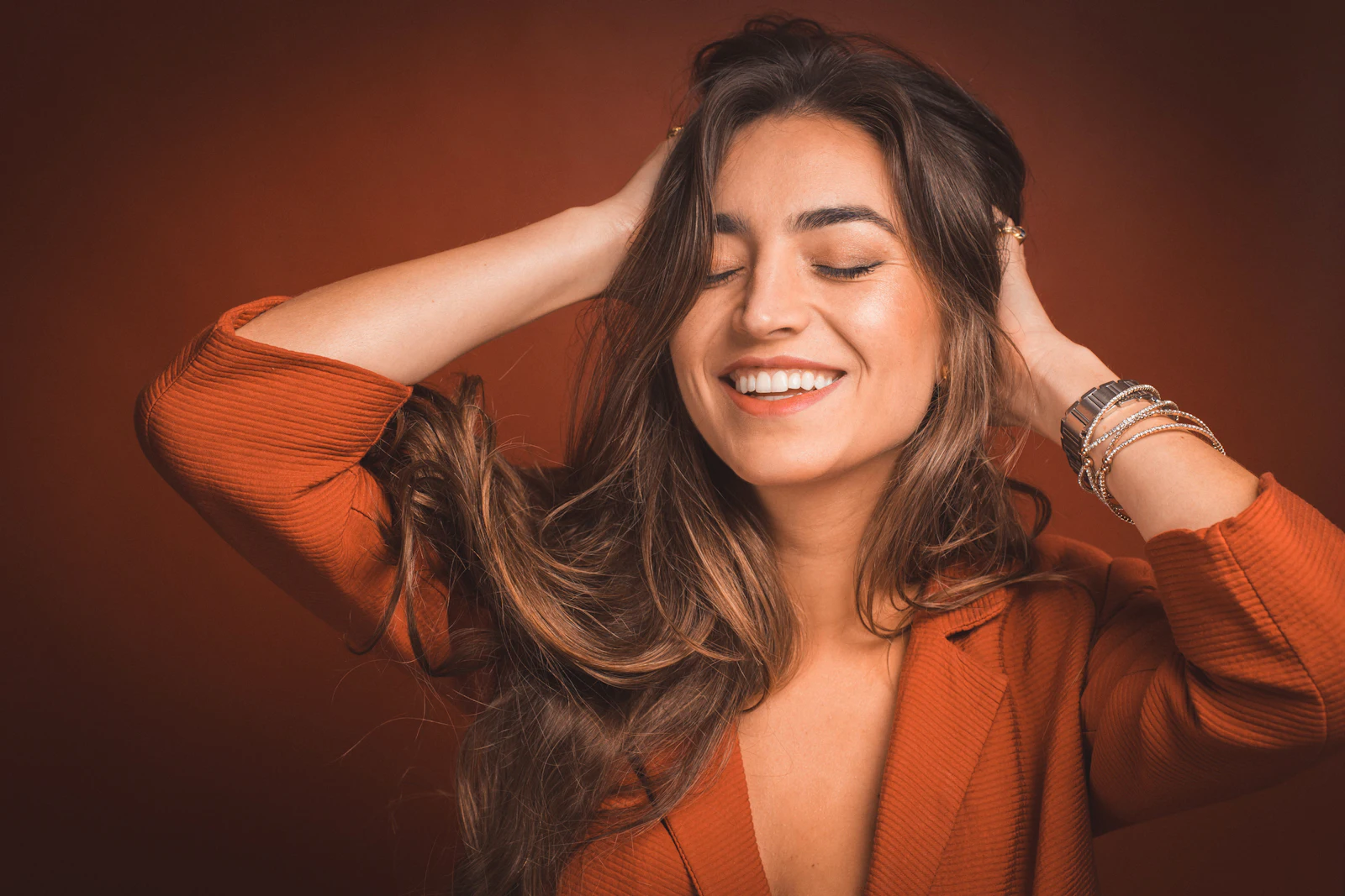 Mulher sorrindo enquanto passa a mão pelos cabelos, de jaqueta cor de terracota